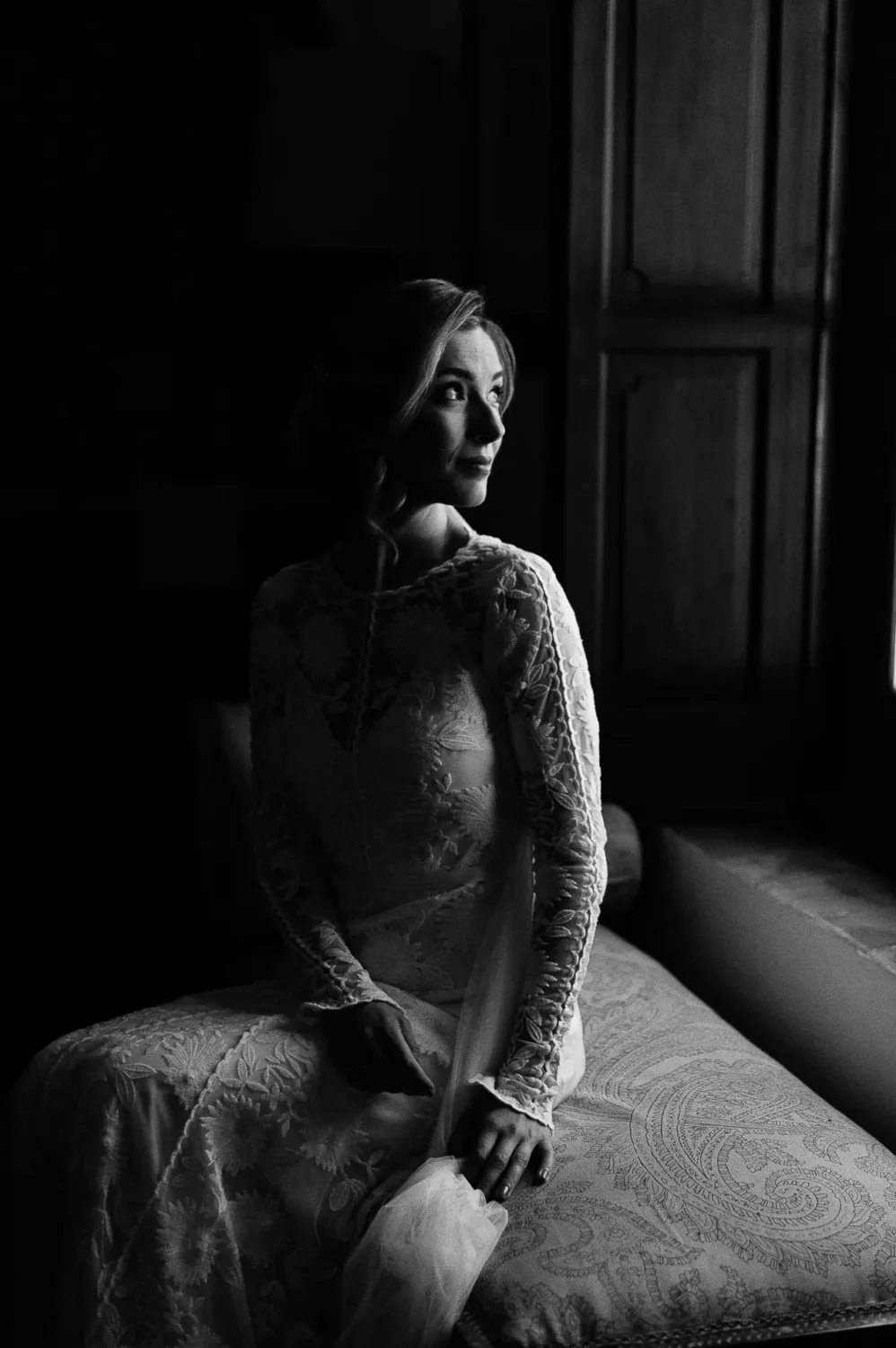 Novia con vestido de encaje sentada junto a una ventana con luz suave, en blanco y negro